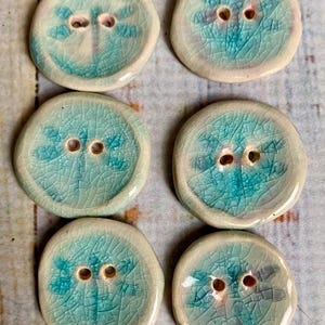 May include: Six handmade ceramic buttons with a dragonfly design. The buttons are round, with a light blue and white color scheme. Each button has two holes for sewing.