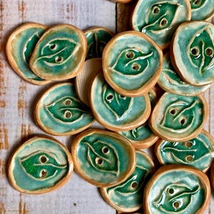 May include: A collection of handmade ceramic buttons. Each button features a leaf design with two holes for sewing. The buttons are teal and green with a brown rim. The buttons are approximately 1 inch in diameter.