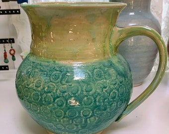 May include: Handmade ceramic pitcher with a textured teal and green exterior. The pitcher has a rounded body, a wide opening, and a curved handle. The top is a light yellow-green color. A second, blue vase is in the background.