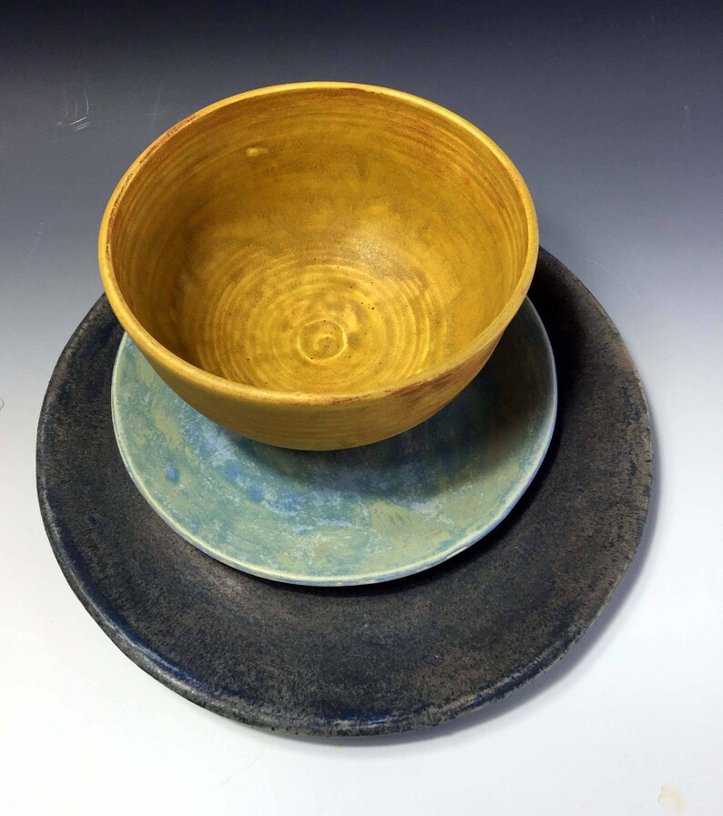 May include: A set of three ceramic dishes in a yellow, blue, and dark gray color scheme. The dishes are stacked on top of each other, with the yellow bowl on top.