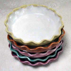 May include: A stack of four ceramic pie plates with scalloped edges. The top plate is yellow, the next is brown, the third is pink, and the bottom is teal.