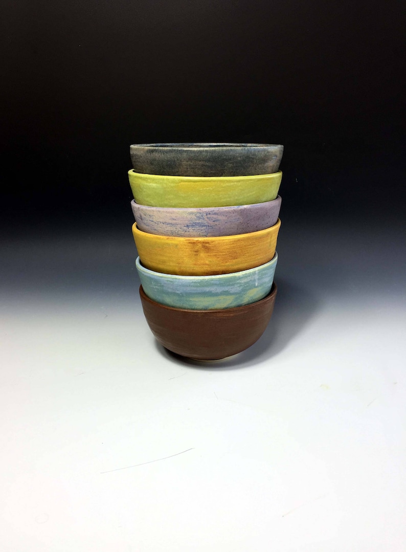 May include: A stack of six ceramic bowls in various colors, including brown, yellow, blue, purple, green, and gray. The bowls are nested inside each other, with the largest bowl at the bottom and the smallest bowl at the top.