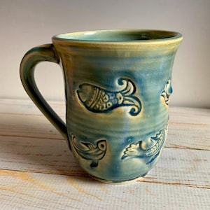 May include: A handmade ceramic mug with a teal and yellow glaze. The mug features embossed fish designs around the exterior. The handle is a matching teal color. The mug is a unique piece of pottery.
