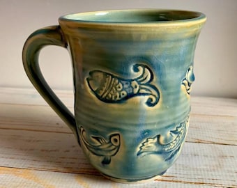 May include: A handmade ceramic mug with a teal and yellow glaze. The mug features embossed fish designs around the exterior. The handle is a matching teal color. The mug is a unique piece of pottery.