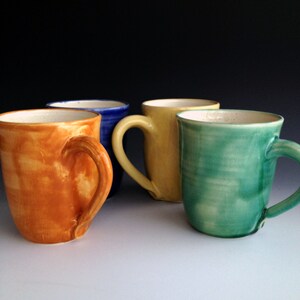 Puede incluir: Cuatro tazas de cerámica en diferentes colores: naranja, azul, amarillo y verde. Las tazas son todas de tamaño y forma similares, con un cuerpo redondeado y un asa.