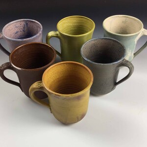 May include: Six ceramic mugs in various colors, including brown, yellow, blue, green, and purple. The mugs are all different shapes and sizes, and they have a rustic, handmade look.