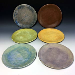 May include: Six ceramic plates in various colors and patterns. The plates are round and have a slightly raised rim. The colors include brown, yellow, green, blue, and purple.