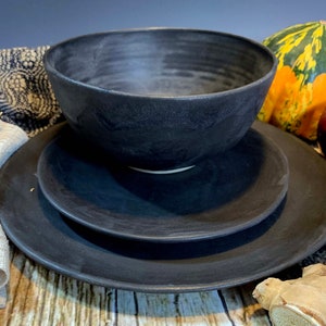 May include: A set of black ceramic dinnerware, including a bowl and two plates. The dishes have a matte finish and are stacked on top of each other.