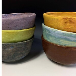 May include: A set of four ceramic bowls stacked on top of each other. The bowls are in shades of purple, green, gray, and brown. The bowls are all oval shaped.