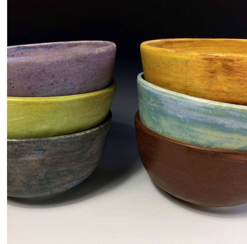 May include: A set of four ceramic bowls stacked on top of each other. The bowls are in shades of purple, green, gray, and brown. The bowls are all oval shaped.