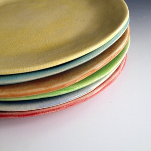 May include: A stack of six ceramic plates in various colors, including yellow, blue, green, orange, and red. The plates are all slightly different sizes and have a crazed glaze.