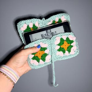 Pode incluir: Uma capa de crochê para leitor de e-books com uma base verde menta clara e três motivos quadrados de granny square. Cada motivo contém um quadrado central amarelo rodeado por fios verde-escuros e rosa-claros. A capa contém um leitor preto.