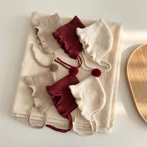 May include: An assortment of baby bonnets in cream, burgundy, and tan. Each bonnet features a ruffled edge and a pom-pom accent. The bonnets are arranged on a cream-colored blanket, with a wooden tray in the background.