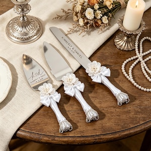 Może przedstawiać: Srebrny zestaw noża i serwera do ciasta z białymi wstążkami i kwiatowymi akcentami. Białe uchwyty mają dekoracyjną srebrną podstawę. Nóż i serwer są grawerowane imionami "KIMBERLY & DANIEL" i datą "16.10.2020".