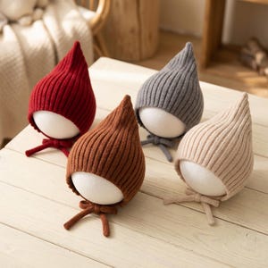 May include: Four knitted baby hats in red, brown, grey, and beige. Each hat has a pointed top and a small bow at the base. The hats are displayed on white, egg-shaped forms, showcasing their shape and texture.