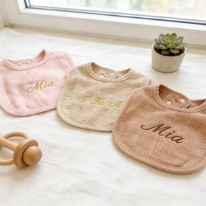 May include: Three personalized baby bibs in pink, beige, and brown, each embroidered with a name in gold thread. A wooden rattle sits nearby. The bibs are made of soft, textured fabric.