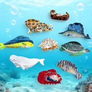 May include: A collection of marine animal-themed hair clips and barrettes. The assortment includes an octopus, a sea turtle, a beluga whale, a fish, a crab, a seashell, and an otter. The clips are in various colors, including blue, yellow, white, and brown.