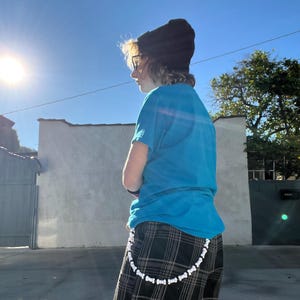 May include: A person wearing a black beanie, blue t-shirt, and a plaid skirt. A white beaded chain hangs from the skirt. The image is taken outside on a sunny day.