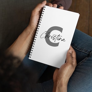 Könnte beinhalten: Ein weißes Spiralnotizbuch, das von einer Person gehalten wird. Das Cover zeigt einen großen grauen Buchstaben "C" mit dem Namen "Christina" in Schreibschrift. Das Notizbuch ist geöffnet und die Seiten sind leer.
