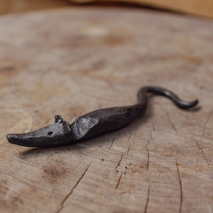 Può includere: Una statuetta di topo in metallo nero con la coda arricciata. La statuetta è realizzata in metallo forgiato e ha un aspetto rustico e artigianale.