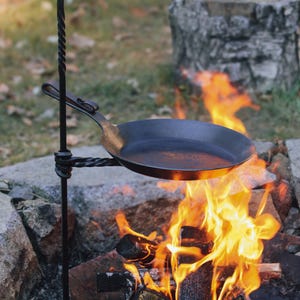 Può includere: Un supporto da cucina in metallo nero con una barra attorcigliata e un gancio nella parte superiore. Una padella in ghisa nera è appesa al supporto sopra un falò.