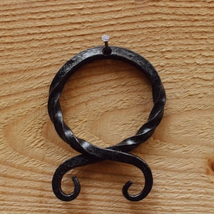May include: Black metal wall hook in the shape of a stylized knot. The hook is attached to a wooden surface with a nail.