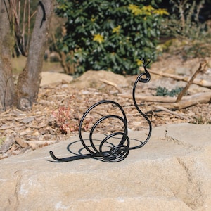 Puede incluir: Escultura de caracol de metal negro con una concha en espiral. El caracol está sentado en una roca marrón claro.