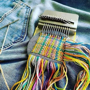 May include: A metal darning loom tool with a series of vertical hooks, currently holding a square patch of woven fabric in vibrant multicoloured threads. The loom is positioned on blue denim fabric, with loose, long, colourful yarn ends hanging down.