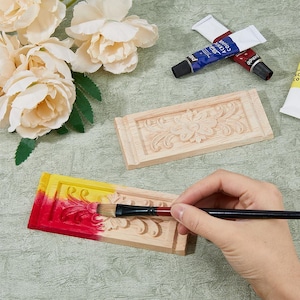 May include: Two rectangular wooden carvings with floral relief patterns, one plain and one partially painted in yellow and red with a paintbrush. Two tubes of acrylic paint are visible, one blue and one red, alongside cream-colored artificial flowers.
