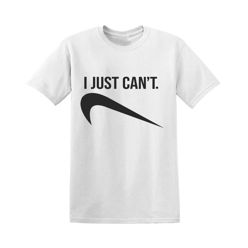 May include: White t-shirt with the text "I JUST CAN'T." above a black swoosh design. The shirt is a crew neck style and appears to be made of a soft material.