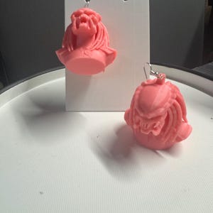 May include: A pair of pink Predator-themed earrings. Each earring features a detailed, sculpted bust of the Predator alien with intricate facial features and dreadlocks. The earrings are displayed on a white earring card.