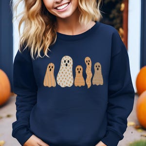 Pode incluir: Camisola azul marinho com cinco aplicações de fantasmas em fila. Os fantasmas têm padrões de riscas, florais e xadrez em tons de laranja e creme. A camisola é de estilo casual, ideal para o outono.