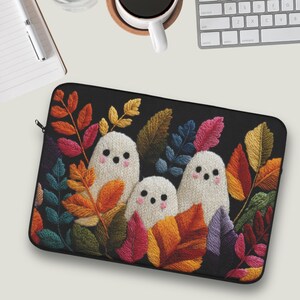 May include: Black laptop sleeve featuring an embroidered design of three white ghosts with black dot eyes and pink blush, surrounded by colorful autumn leaves in shades of orange, red, yellow, and green. The sleeve has a black zipper.