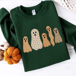 May include: A dark green sweatshirt with an appliqué design of five ghosts. The ghosts are patterned with stripes, floral, and plaid in orange, cream, and brown. The sweatshirt is displayed on a white surface with decorative pumpkins.