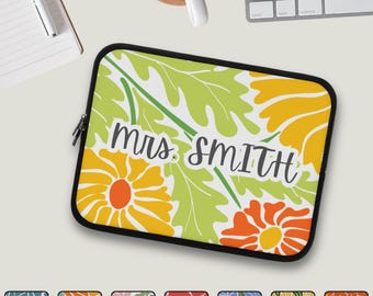 May include: A black-edged laptop sleeve featuring a floral design with green leaves and orange and yellow flowers. The text "Mrs. Smith" is written in a cursive font across the center of the sleeve.