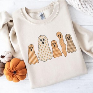 Op de afbeelding: Crèmekleurige sweatshirt met een ontwerp van vijf spookfiguren. Elke geest heeft een uniek patroon, waaronder strepen, bloemen en ruiten, in tinten oranje, bruin en crème. Het ontwerp is gecentreerd op de voorkant van de sweatshirt.
