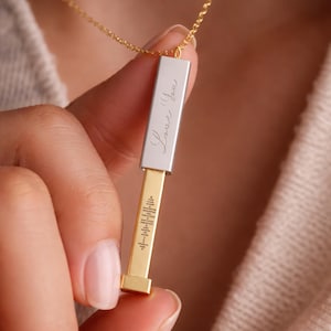 May include: A gold pendant necklace with a rectangular bar design. The top section is silver with the engraved text "Love You". The bottom section has a gold finish with a sound wave pattern. The necklace is held by a person.