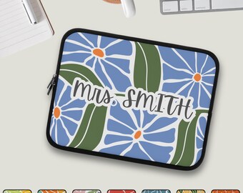 May include: A black laptop sleeve featuring a floral design with blue flowers, green leaves, and orange centers. The text "Mrs. Smith" is displayed in a handwritten style. The sleeve has a black zipper and trim.