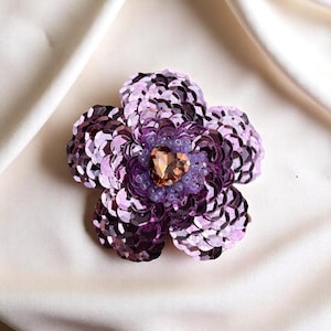 Könnte beinhalten: Eine violette Blumenbrosche aus Pailletten mit einem großen, klaren Strassstein in der Mitte.