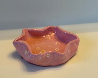 May include: A small, pink ceramic dish with a scalloped edge and a white floral pattern.