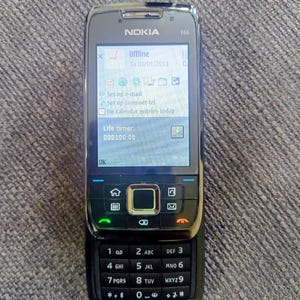 Könnte beinhalten: Ein schwarzes Nokia E66 Mobiltelefon mit einem Bildschirm, der "Offline" und das Datum "Sa 01/01/2011" anzeigt. Das Telefon hat eine Tastatur mit Zahlen und Buchstaben sowie verschiedene Funktionstasten. Das Telefon liegt auf einer grau strukturierten Oberfläche.