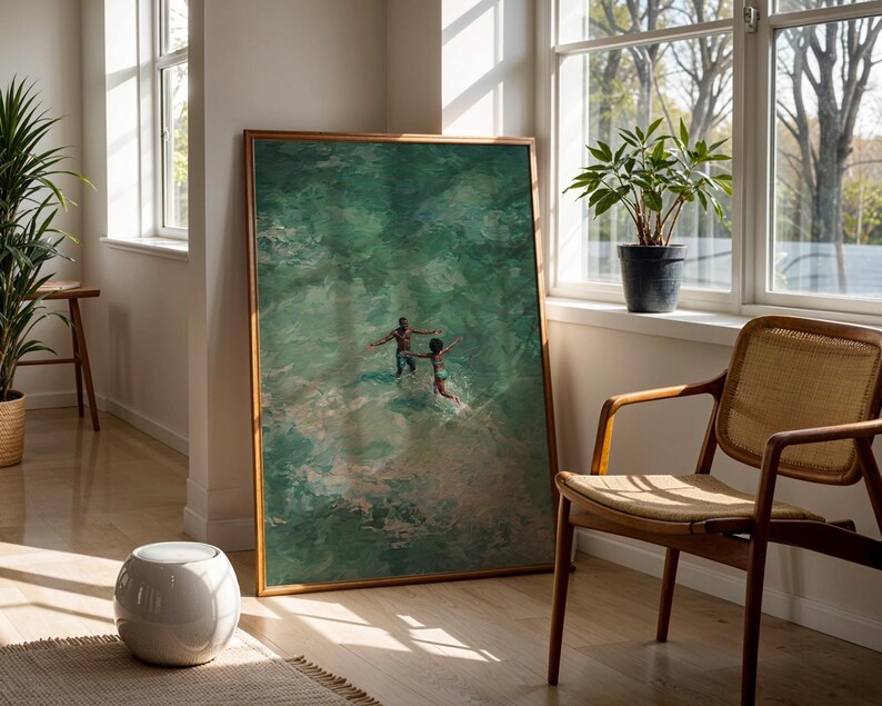 May include: A framed art print depicts two figures in turquoise water, arms outstretched. The artwork is displayed in a well-lit room with a wooden chair, a potted plant, and a white ceramic stool. The frame is a warm, natural wood tone.