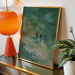 May include: A framed art print with a gold frame depicts two figures in the ocean. An orange lamp with a ribbed shade and a conical base sits on a glass-topped table. A potted plant with green and yellow leaves is on the right.