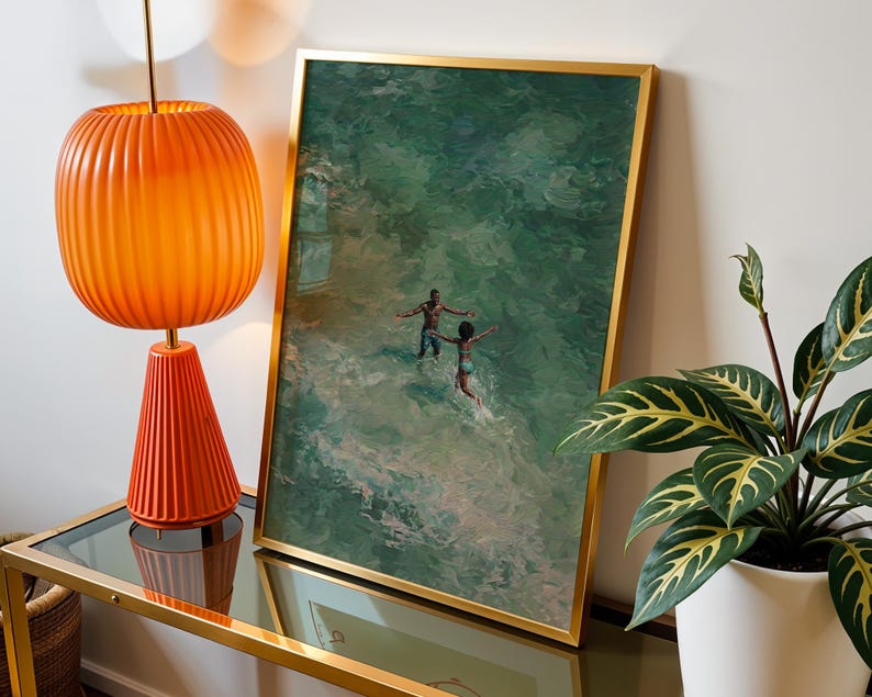 May include: A framed art print with a gold frame depicts two figures in the ocean. An orange lamp with a ribbed shade and a conical base sits on a glass-topped table. A potted plant with green and yellow leaves is on the right.