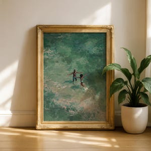 May include: A framed art print featuring a painting of two people in turquoise water, arms outstretched. The artwork is in a gold ornate frame, leaning against a white wall. A potted plant is to the right, with a wooden floor.