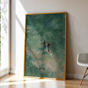 May include: A framed art print depicts two figures in a body of water, with a gold frame. The water is a mix of green and white hues. The artwork is leaning against a white wall next to a window and a modern chair.