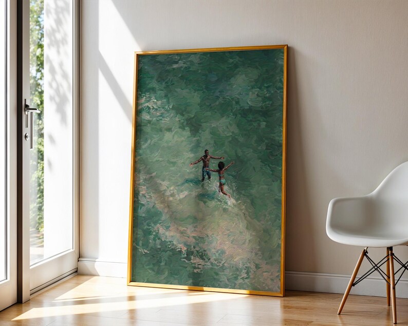 May include: A framed art print depicts two figures in a body of water, with a gold frame. The water is a mix of green and white hues. The artwork is leaning against a white wall next to a window and a modern chair.
