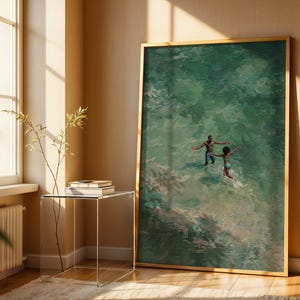 May include: A framed art print depicts two figures in turquoise water, arms outstretched. The artwork, with a gold frame, leans against a beige wall. A clear side table with books and a plant sits near a window with sunlight.