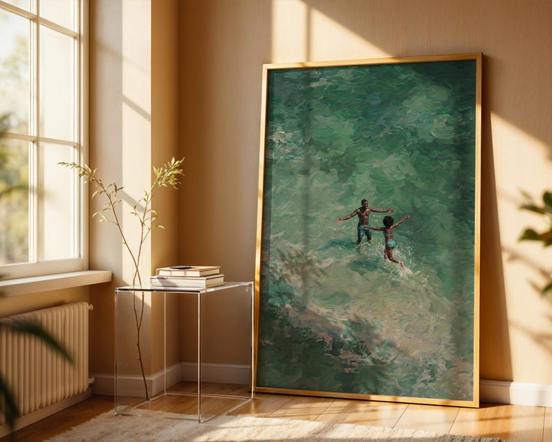 May include: A framed art print depicts two figures in turquoise water, arms outstretched. The artwork, with a gold frame, leans against a beige wall. A clear side table with books and a plant sits near a window with sunlight.