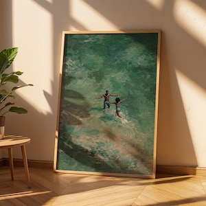 May include: A framed art print depicts two people in a turquoise ocean. The artwork has a warm, natural wood frame. The figures are in the water with their arms outstretched, creating a sense of joy and movement. The art is leaning against a wall.